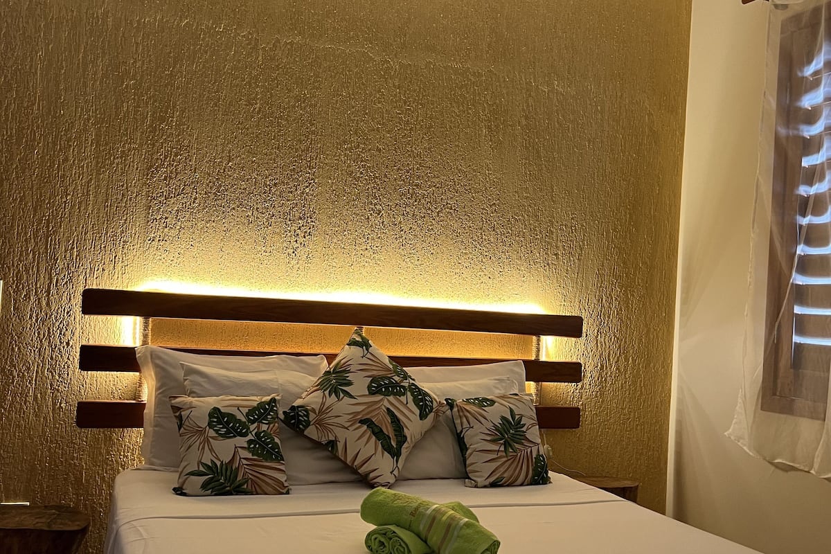 Un dormitorio con cama, cabecera de madera y colcha con estampado de hojas tropicales.