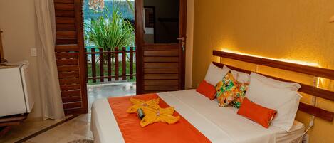 A hotel room with a bed, a wooden headboard, a small table, and a view of a garden through an open door.