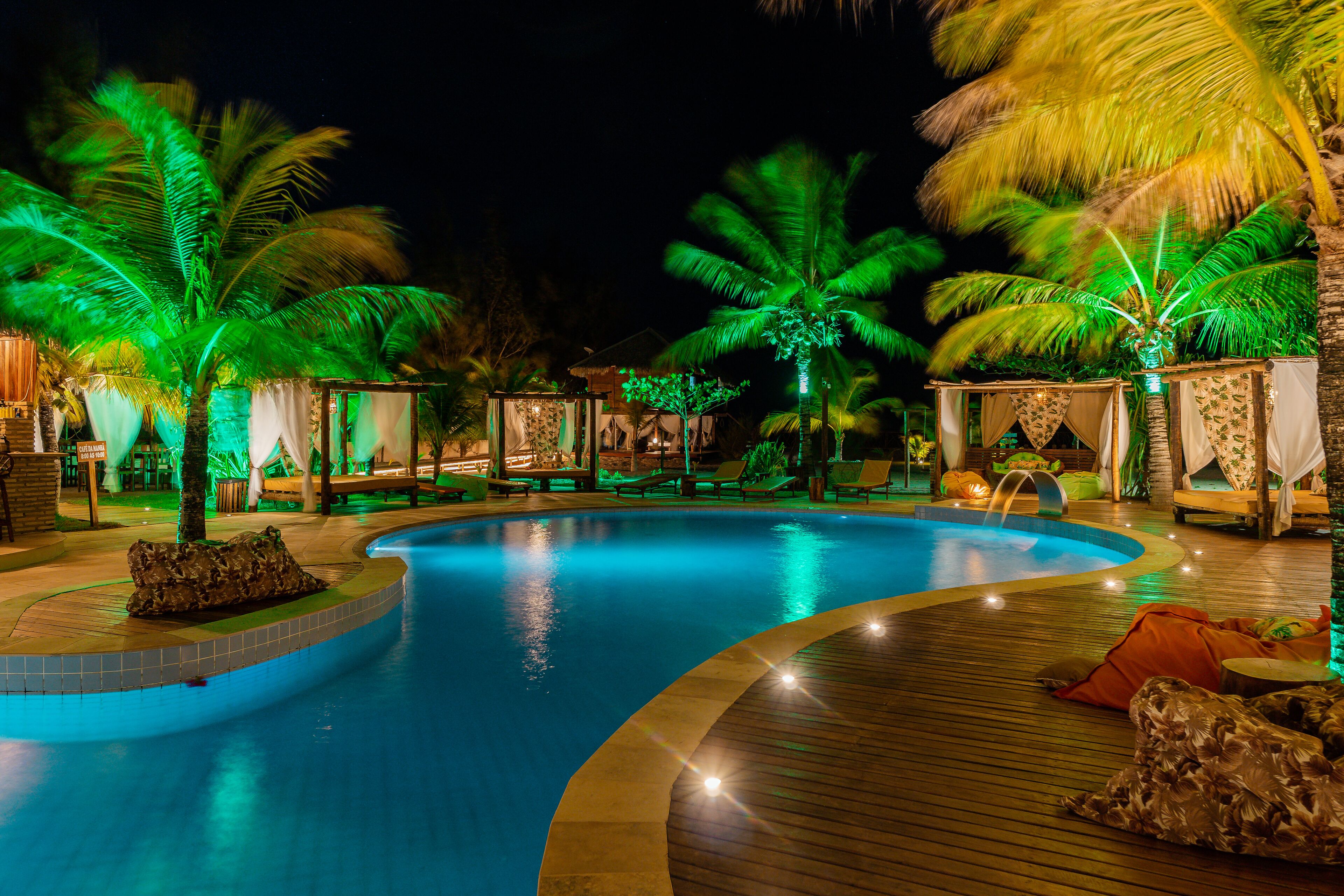 Un área de piscina nocturna con piscina curvada, palmeras y sillas de descanso.