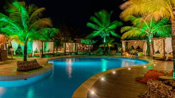 Un área de piscina nocturna con piscina curvada, palmeras y sillas de descanso.