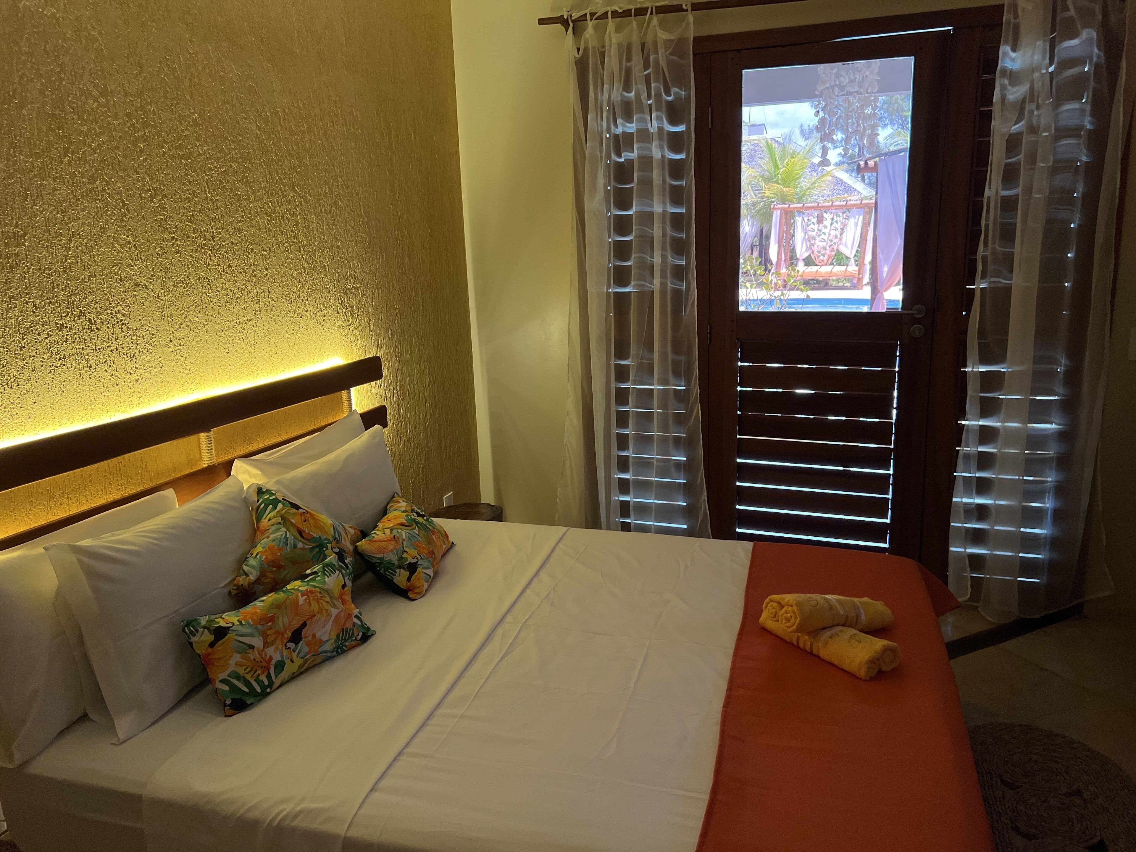 Una cama bien hecha con almohadas blancas y coloridas, una manta roja y una toalla enrollada al lado.