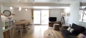 Un salon moderne avec une table à manger en bois, des chaises transparentes, un canapé noir et un téléviseur posé sur un meuble.