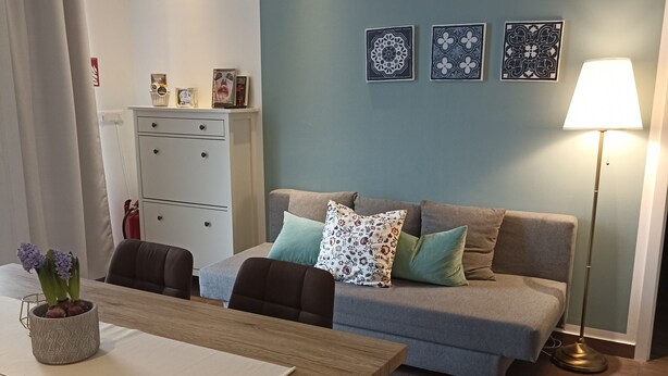 A modern living room with a wooden dining table, a sofa with patterned cushions, and a floor lamp.