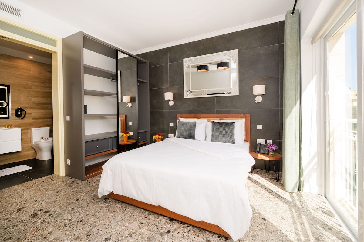 Une chambre d ’hôtel moderne dotée d’un grand lit, d’une armoire intégrée, d’une salle de bain avec des toilettes et un lavabo, et d’une grande fenêtre laissant entrer la lumière naturelle.