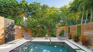 Une piscine rectangulaire, agrémentée de deux chaises longues et d’une petite table, le tout entouré d’une clôture en bois et d’une végétation luxuriante.