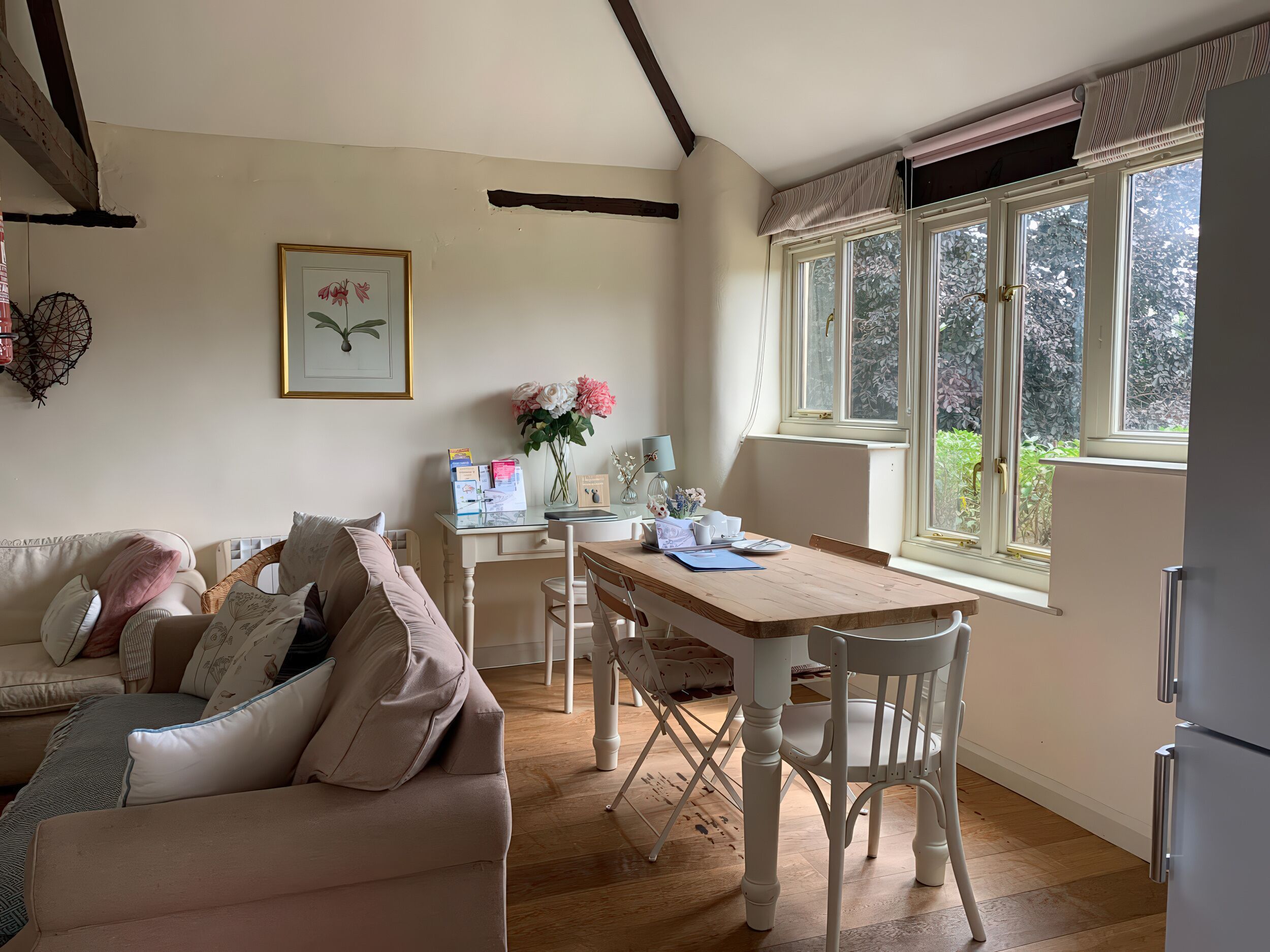 A cozy living room with a wooden dining table, white chairs, a beige sofa, and a large window with a view of greenery.