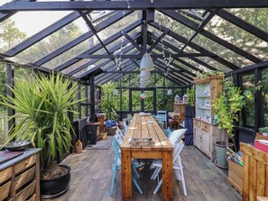 A greenhouse with a wooden table and chairs, surrounded by potted plants and a wooden cabinet with shelves. - The Bull Pen (Colyton)