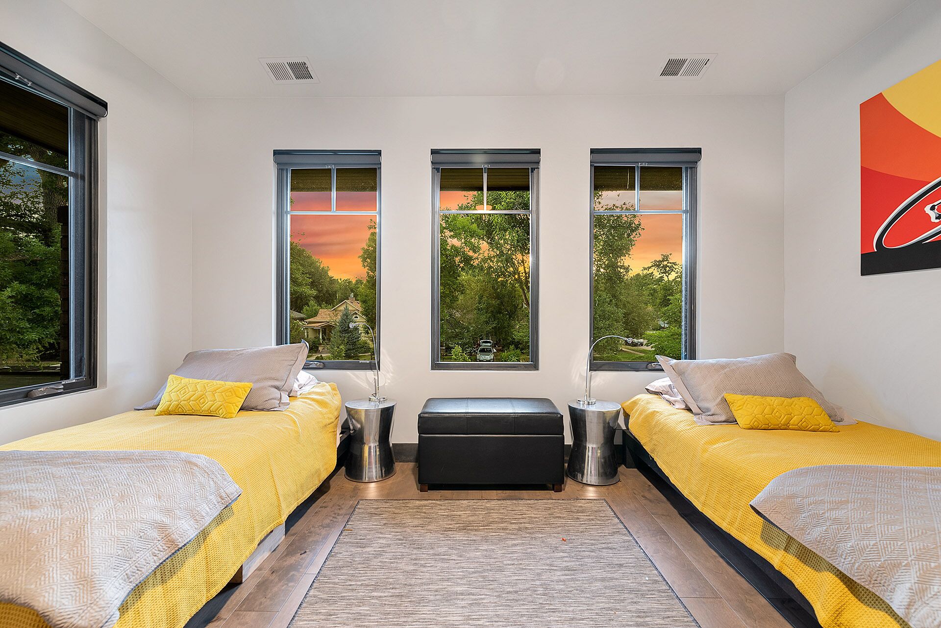 Un dormitorio moderno con dos camas, un banco central y ventanas amplias que ofrecen vista a árboles y un atardecer.