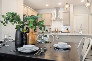 Une table à manger dressée avec des assiettes, des verres et des couverts, entourée de plantes en pot et d’une bouteille de vin.