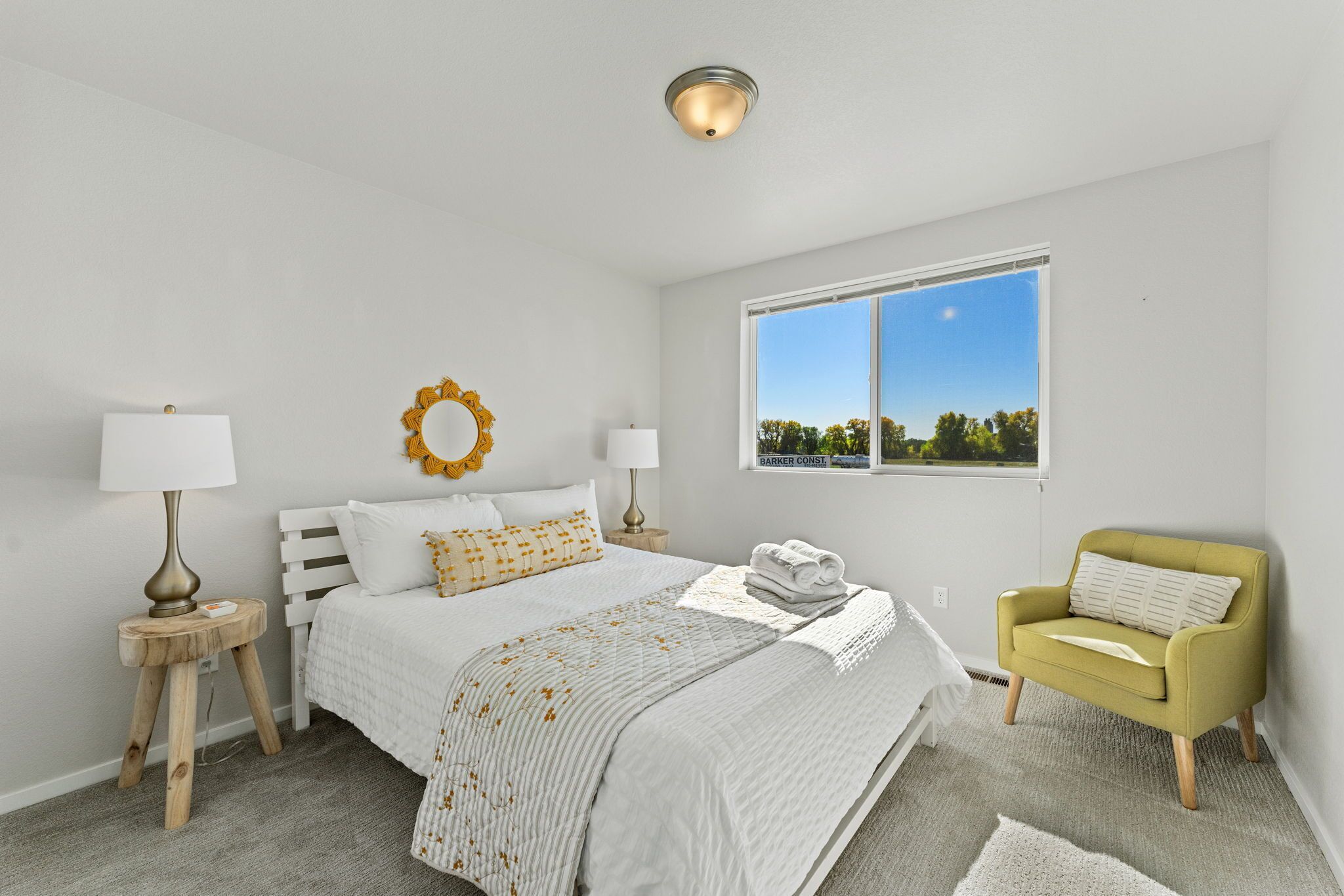 A bedroom with a bed, bedside lamps, a window with a view, a yellow armchair, and a small wooden stool.