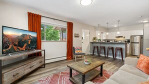 Un salon moderne avec un téléviseur, un canapé et une table basse, une cuisine avec des tabourets de bar et une grande fenêtre avec des rideaux.