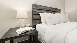 Un lit bien fait, avec une literie blanche, une table de chevet avec une lampe et un livre, et une étagère murale avec une tasse.
