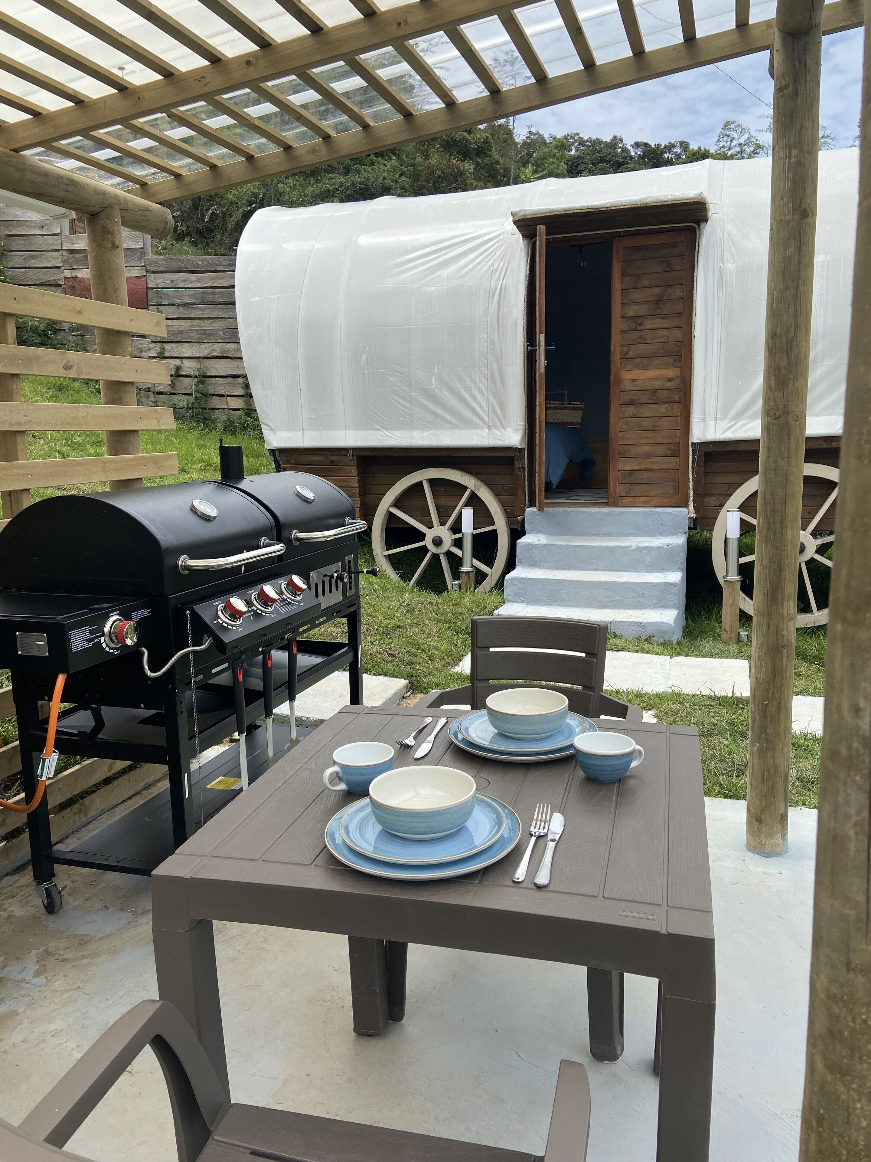 Un área de comedor al aire libre con una mesa preparada para cuatro, una parrilla y un carro cubierto al fondo.
