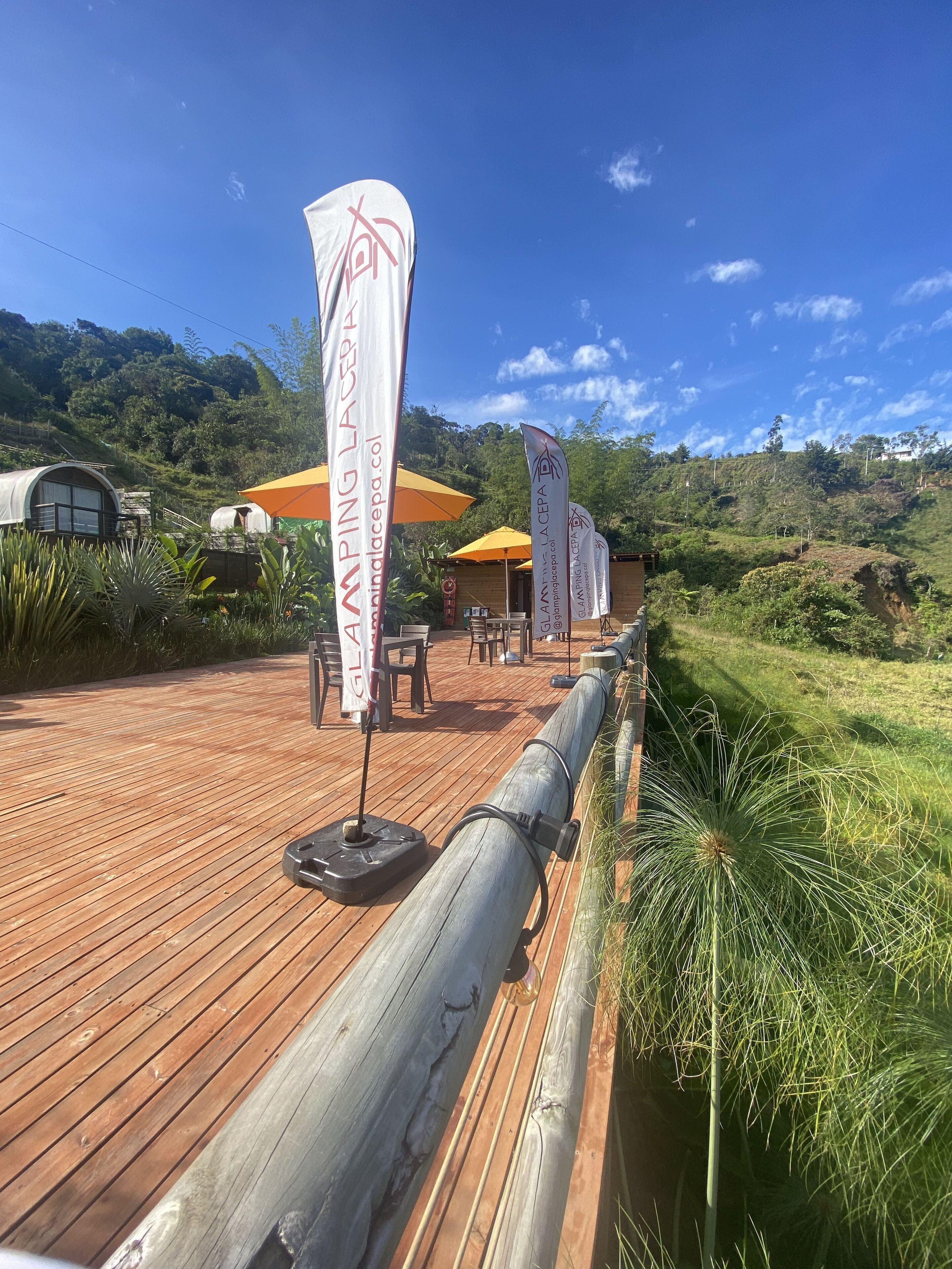 A wooden deck with outdoor seating and flags, surrounded by lush greenery and hills.