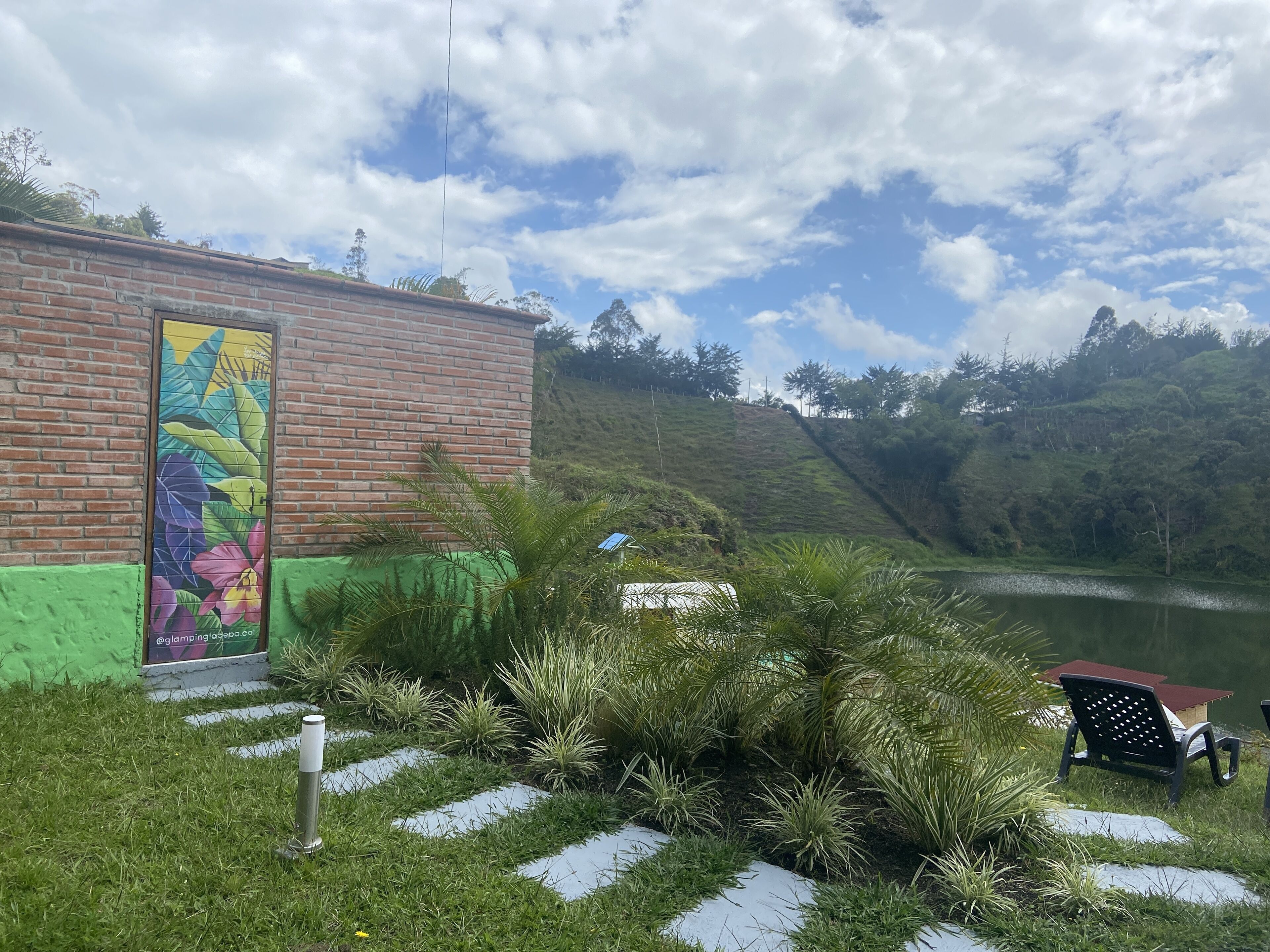A building with a colorful mural, a grassy area with plants, and a lake in the background.