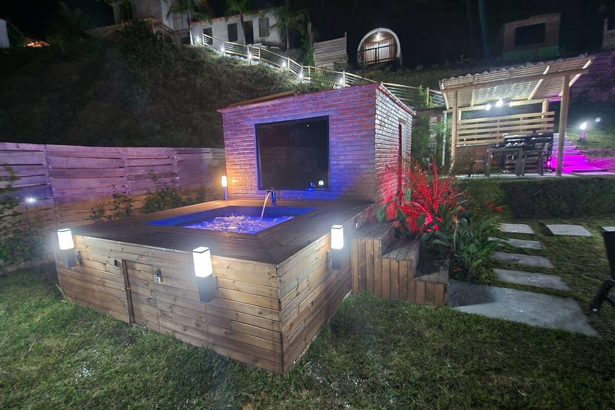 A wooden hot tub with a built-in seating area, surrounded by a wooden deck and a small brick structure with a window.