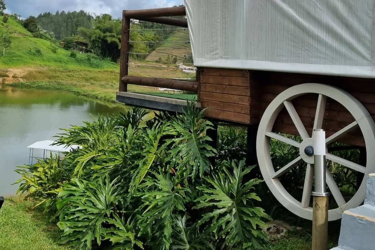 A covered outdoor seating area with a wooden bench and a large wheel structure, surrounded by lush greenery and a body of water.