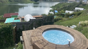 A hot tub with a wooden deck, surrounded by lush greenery and overlooking a calm lake with houses in the background. - Glamping La Cepa GUATAPÉ (El Peñol)