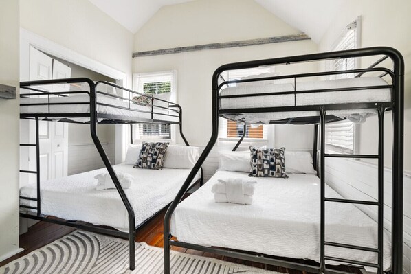 A room with two bunk beds, white bedding, patterned pillows, a wooden floor, and a window with blinds.