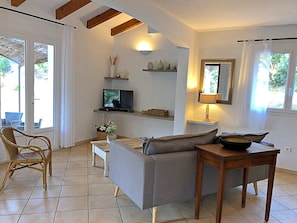 A living room with a grey sofa, a wooden table, a TV on a shelf, and a window with curtains. - 2 3* POOL VILLAS WITH PANORAMIC VIEWS BETWEEN THE SEA AND THE MOUNTAINS (Porto-Vecchio)