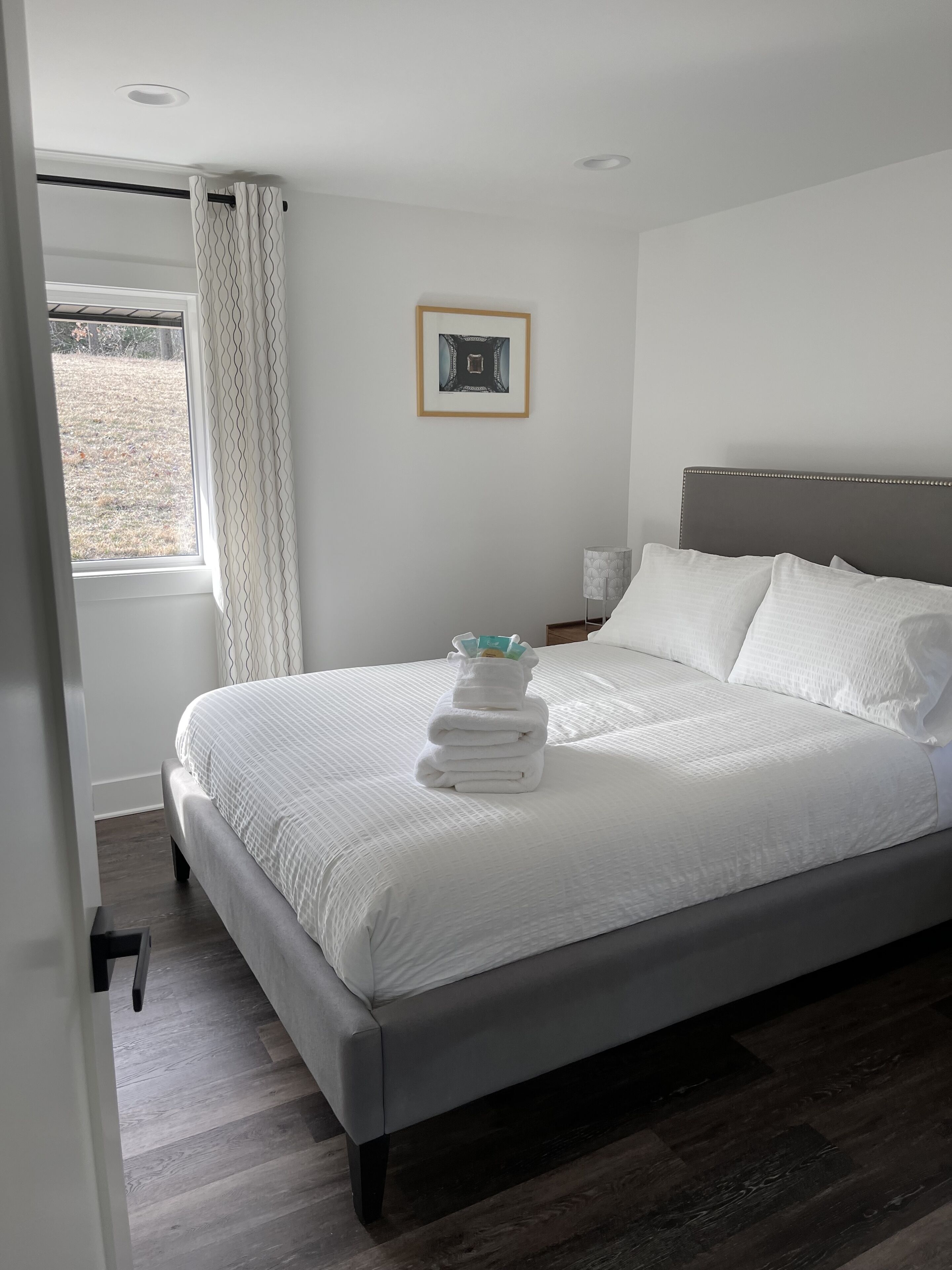 A neatly made bed with white linens and a stack of folded towels, a framed picture on the wall, and a window with curtains.