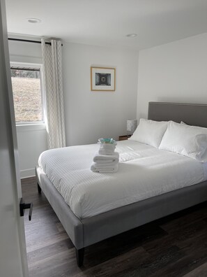 Un lit bien fait, avec du linge de lit blanc et une pile de serviettes pliées, un tableau encadré au mur et une fenêtre avec des rideaux.