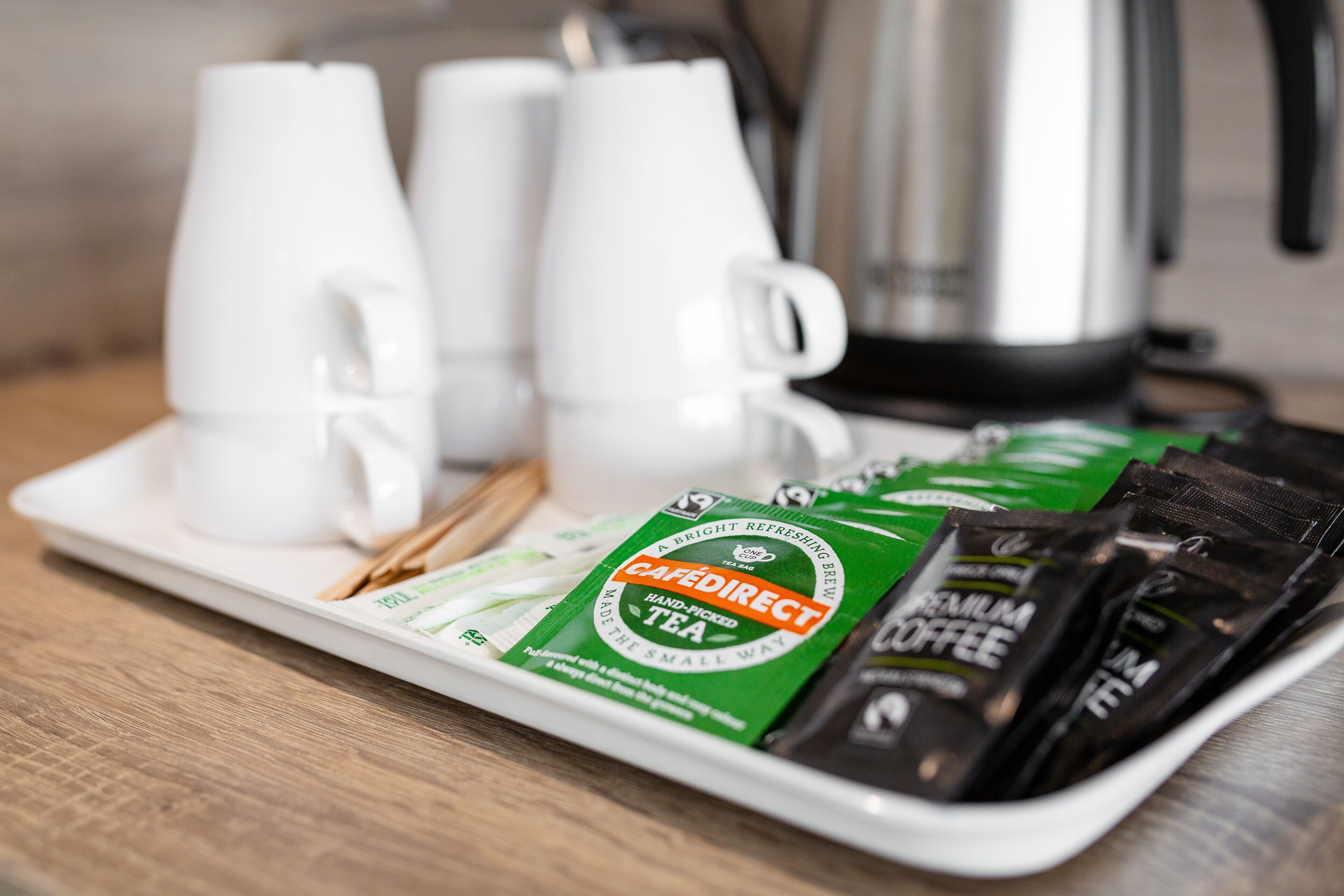 A tray with coffee and tea packets, a white coffee pot, and two white cups on a wooden table.