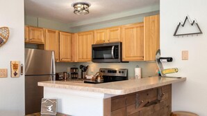 A kitchen with wooden cabinets, a stainless steel refrigerator, a microwave, an oven, and a coffee machine. There is a wooden island with stools and a small table with a sign. - Waterside West A303 Ideal and modern 1 bedroom - open floor plan and mt views (Fraser)