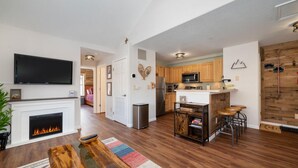 A spacious living room with a fireplace, a television, and a kitchen area with wooden cabinets and a microwave. - Waterside West A303 Ideal and modern 1 bedroom - open floor plan and mt views (Fraser)