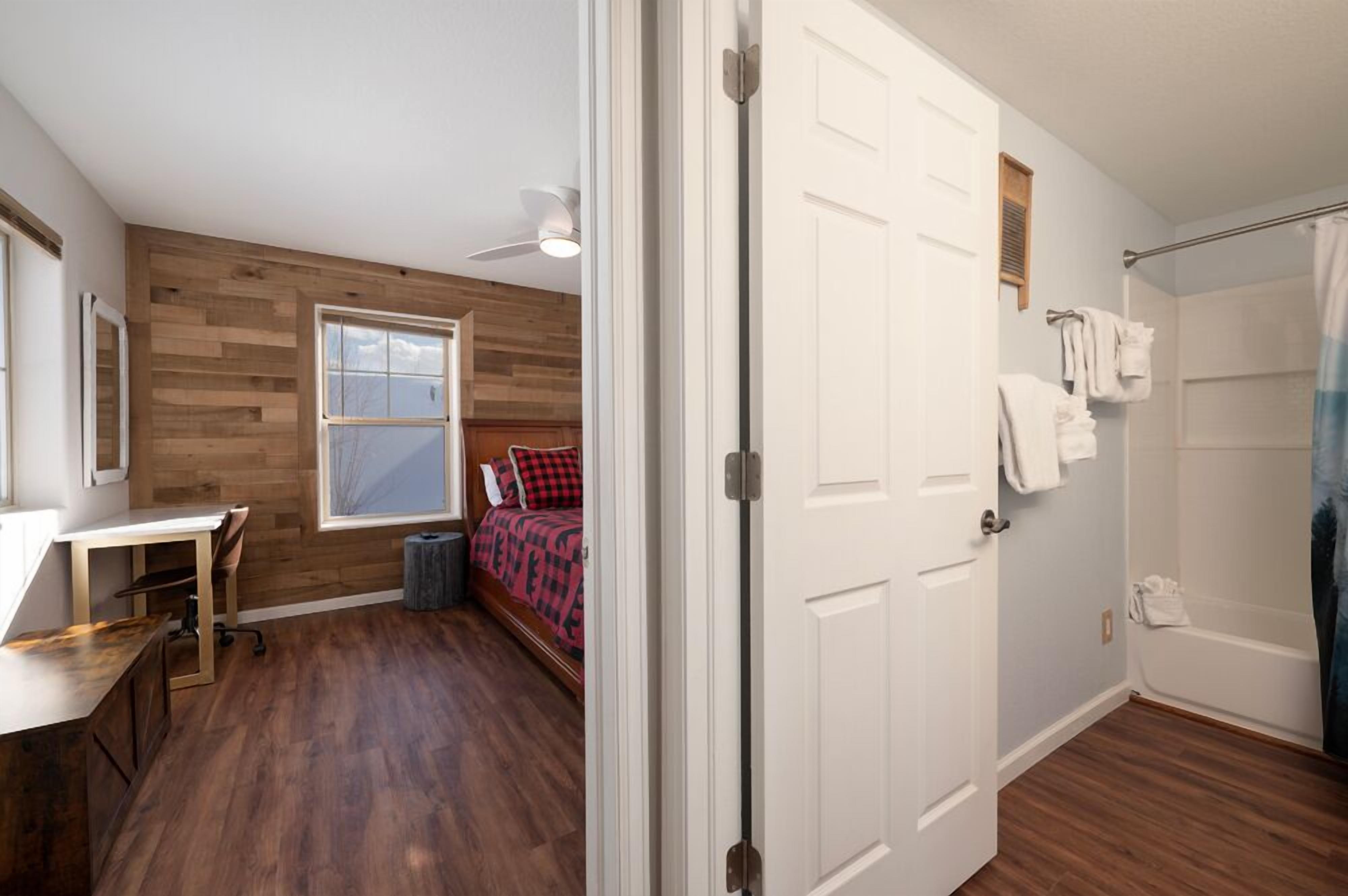 A bedroom with a wooden floor, a desk, a bed with a red and black checkered blanket, a window, and a white door.