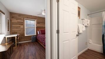 A bedroom with a wooden floor, a desk, a bed with a red and black checkered blanket, a window, and a white door.