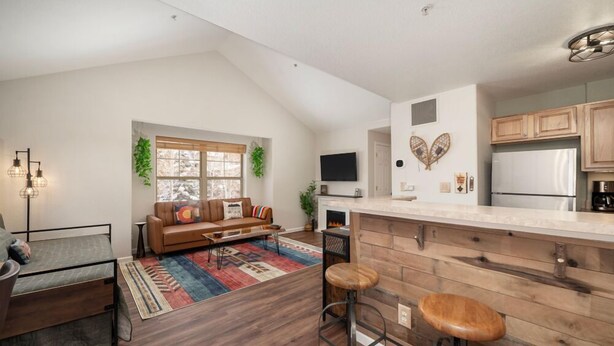 A modern kitchen with wooden cabinets, a built-in island with stools, a flat-screen TV above the island, and a fireplace. - Waterside West A303 Ideal and modern 1 bedroom - open floor plan and mt views (Fraser)