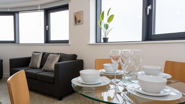 Un salon moderne avec une table à manger en verre pour quatre personnes, un canapé noir et une plante en pot près de la fenêtre.