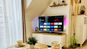 A modern living room with a TV mounted on a sloped ceiling, a wooden coffee table, a round table, and a woven rug. - The roofs of Nazado (2/4 people) (Erquy)