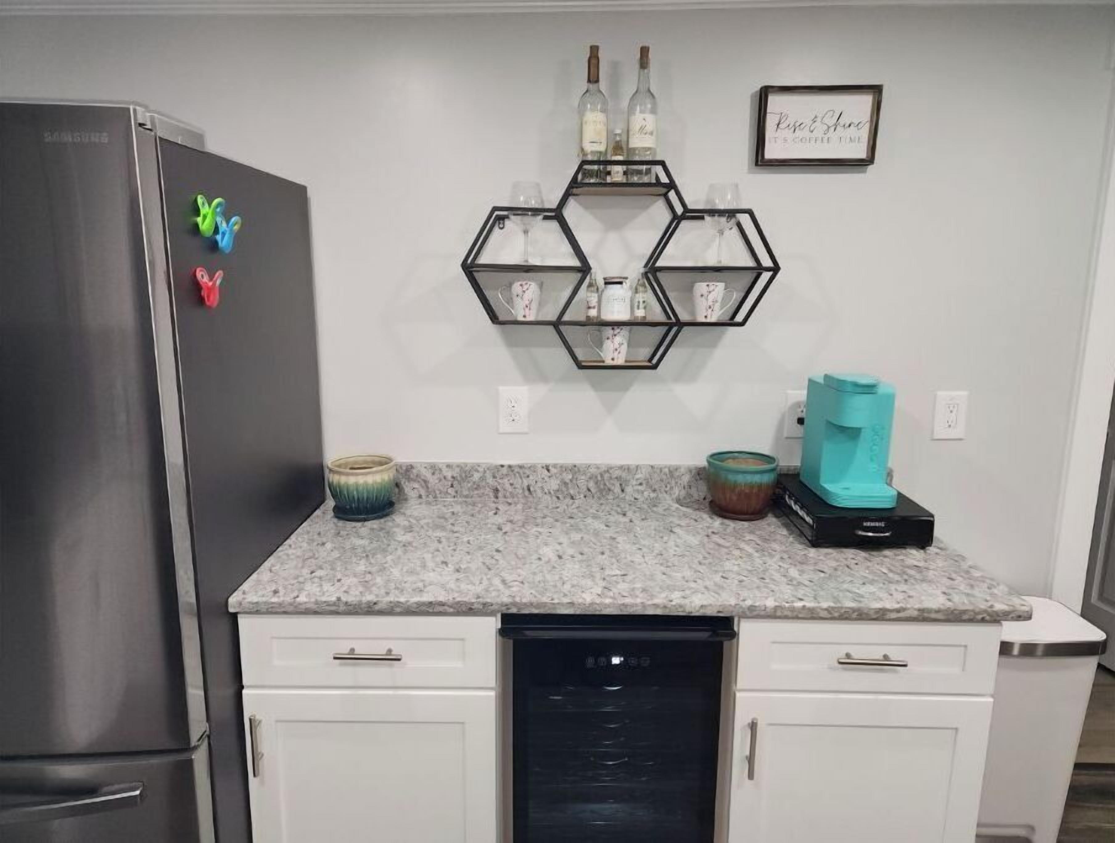 A kitchen with a stainless steel refrigerator, a dishwasher, and a countertop with a hexagonal shelf holding wine bottles and glasses.