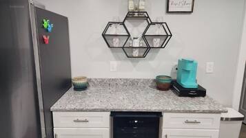 A kitchen with a stainless steel refrigerator, a dishwasher, and a countertop with a hexagonal shelf holding wine bottles and glasses.