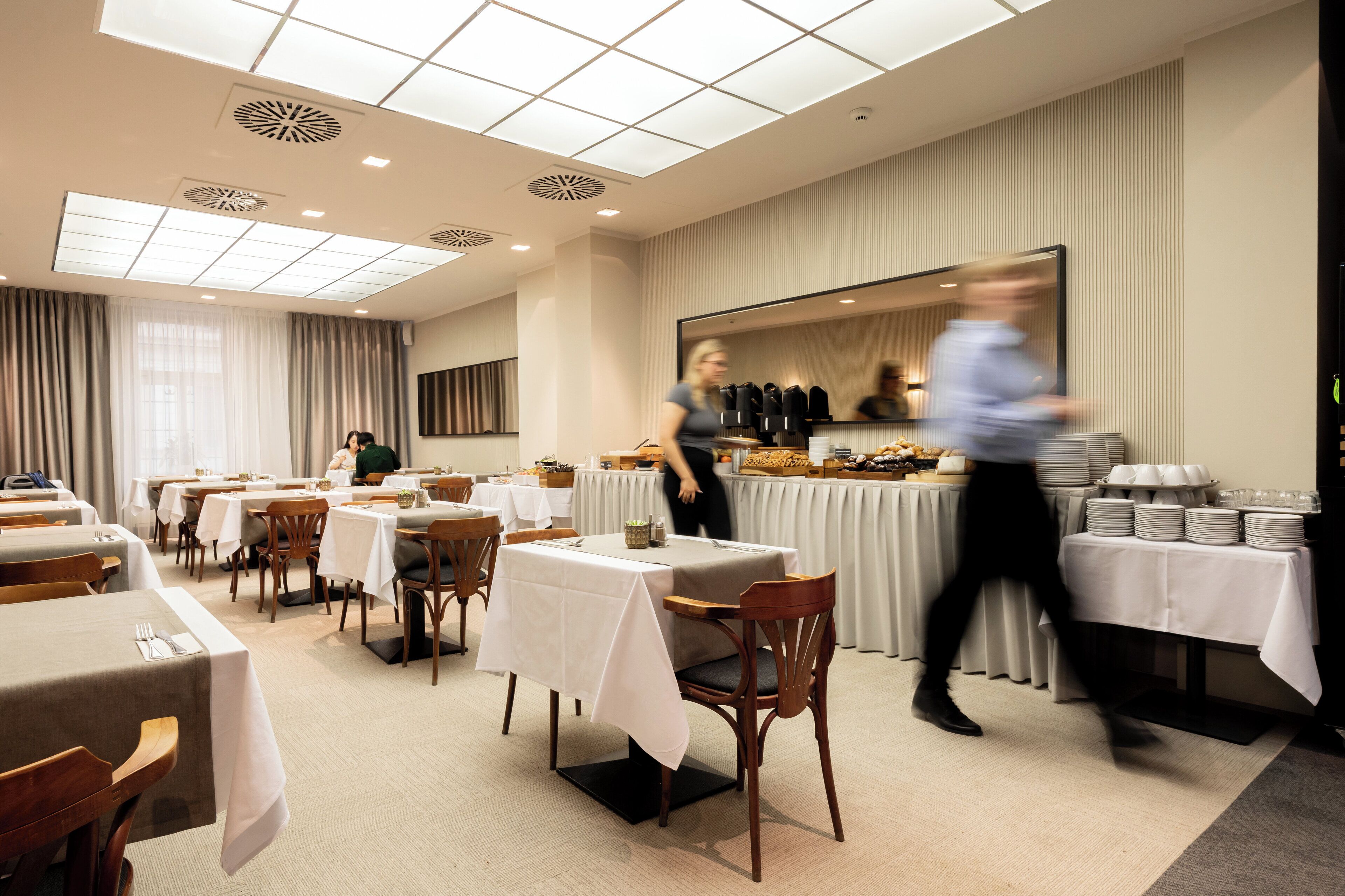 A hotel dining area with tables set for guests, a buffet line, and a large mirror reflecting the space.