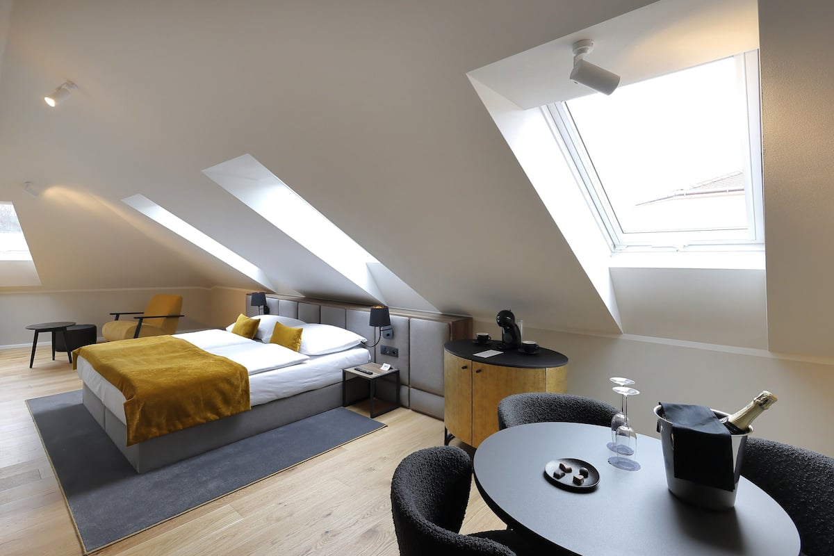 A modern hotel room with a bed, a small desk, a chair, a table, and a window.