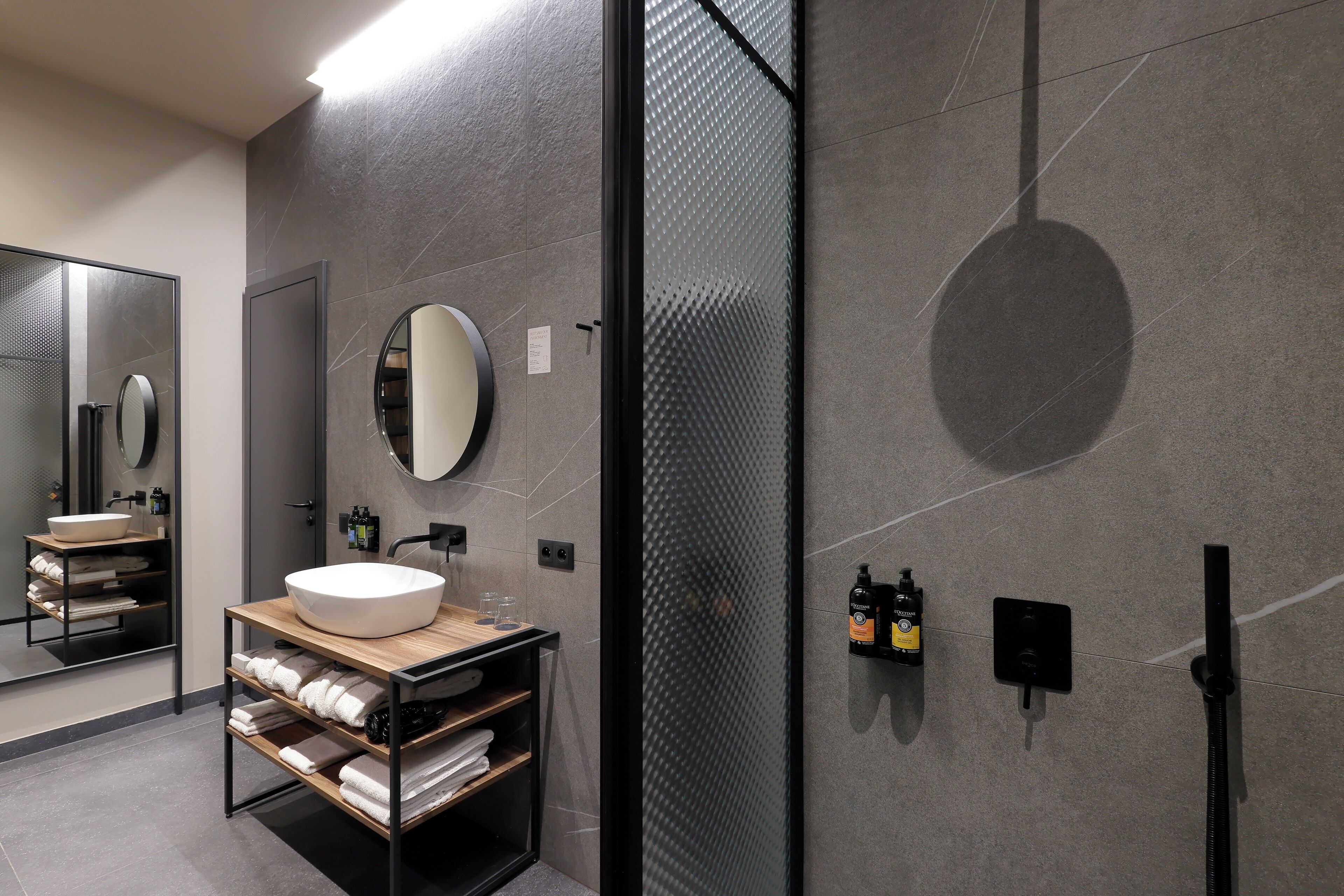 Un baño moderno con un espejo grande, un espejo redondo, un estante de madera con toallas y un lavabo sobre encimera.