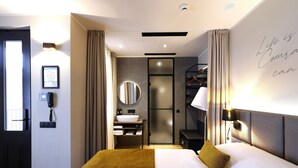 Une chambre d’hôtel moderne avec un grand lit, une table de chevet, un téléphone fixé au mur, une salle de bain avec un lavabo et un miroir, et une citation inspirante sur le mur.