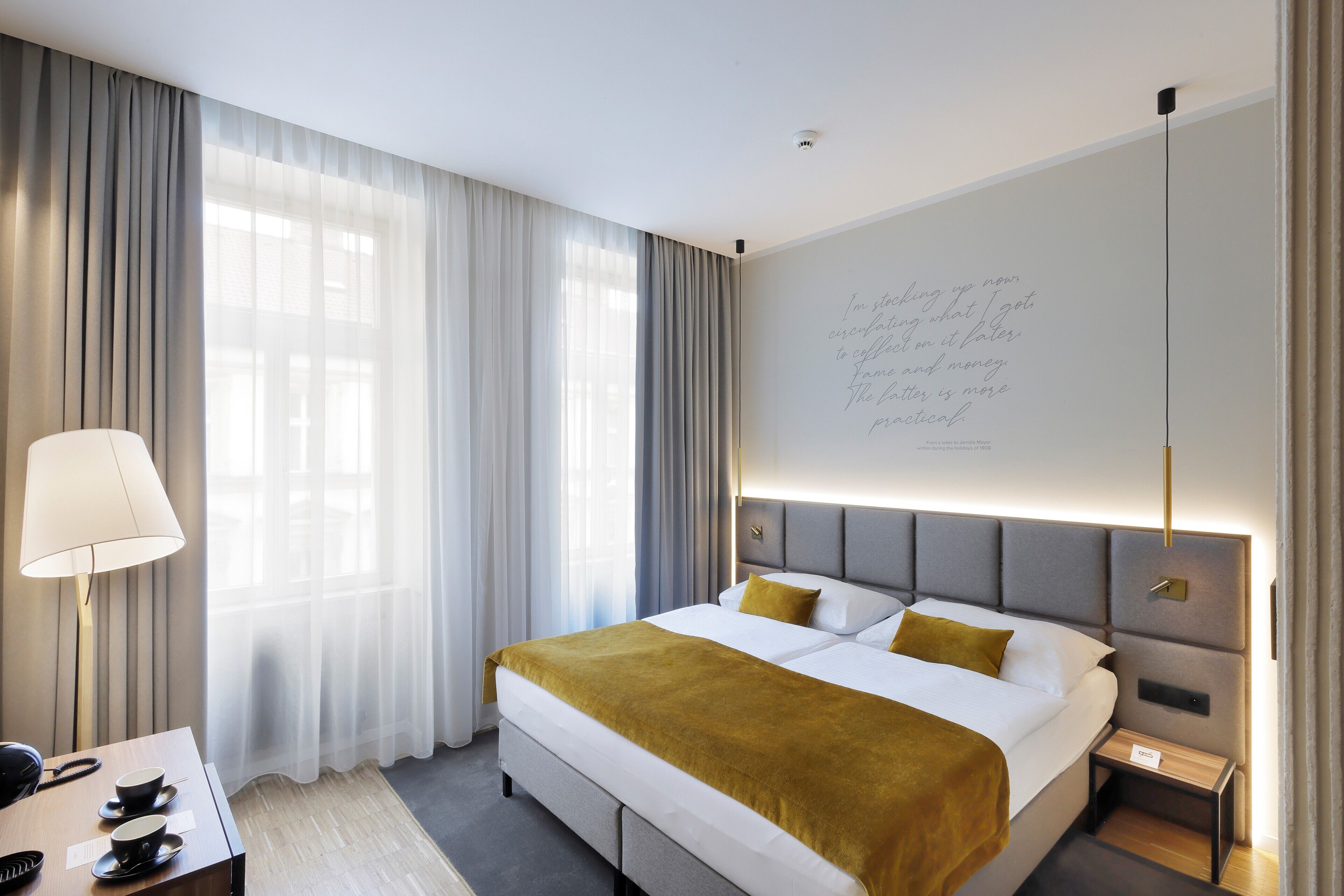 A modern hotel room with a large bed, a bedside table with a lamp, and a wall with a motivational quote.