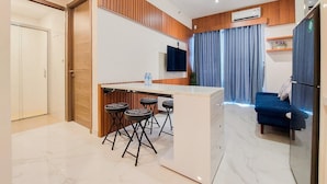 A modern kitchen with a central island, bar stools, a flat-screen TV, and a blue sofa in a living area. - Fully Furnished And Modern 2Br Apartment At Sky House Bsd (Pagedangan)