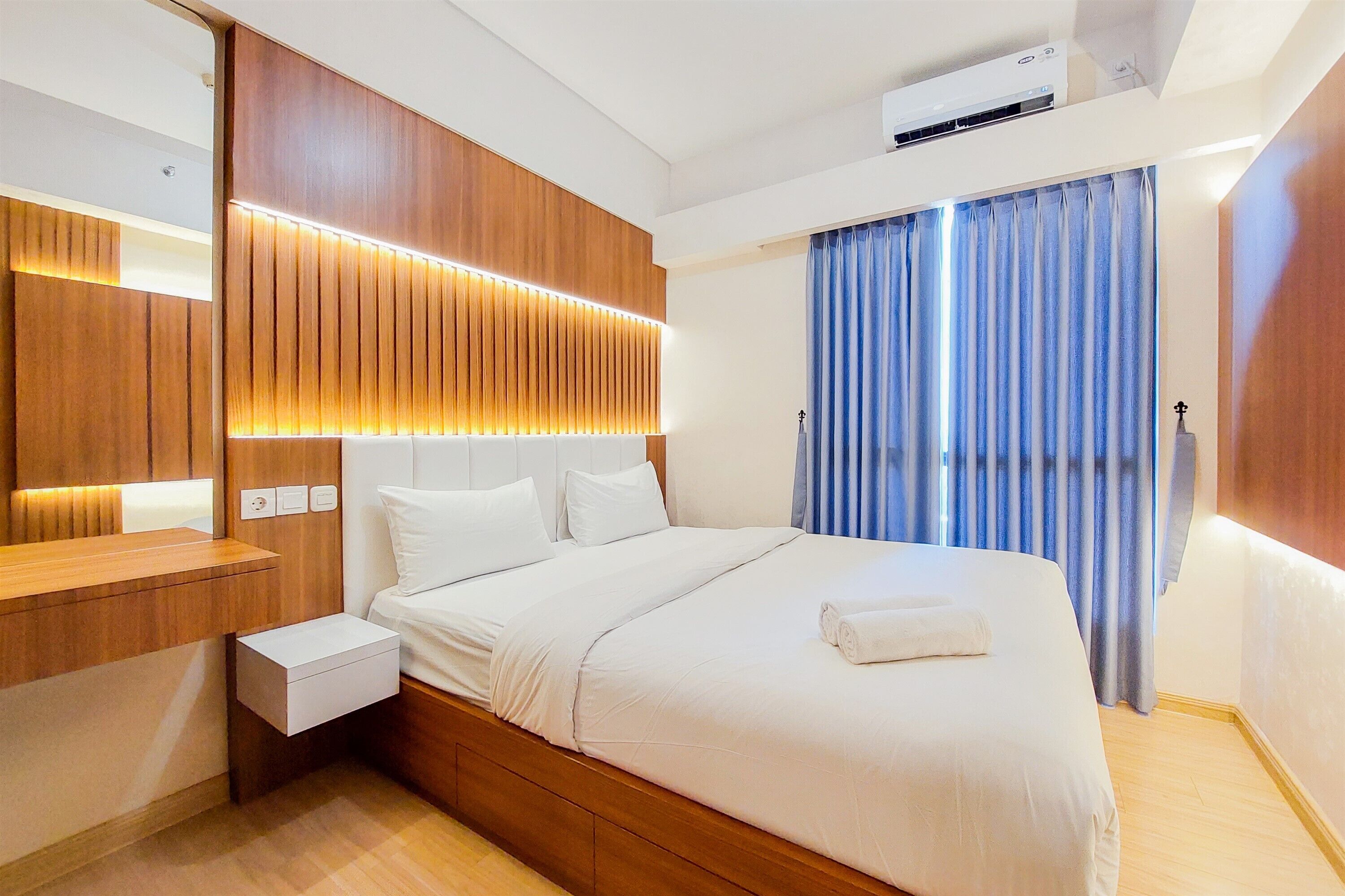 A modern hotel room with a wooden headboard, a large bed with white linens, a bedside table, and a window with blue curtains.