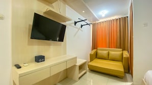 Ein modernes Hotelzimmer mit einem Wandfernseher, einem beigefarbenen Sofa und einem kleinen Holztisch mit Fernbedienung und einer Schachtel.