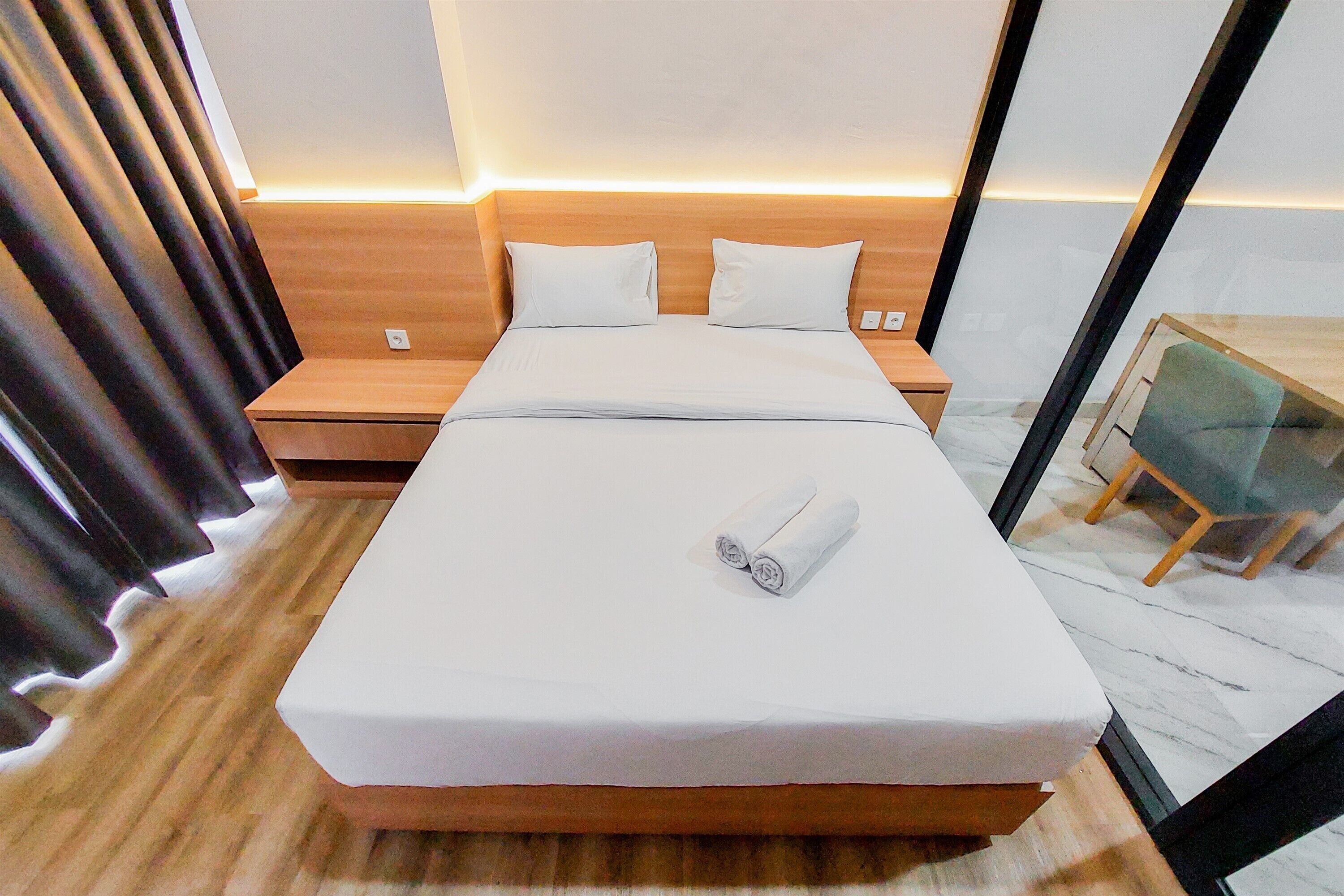 A neatly made bed with white linens and a rolled towel, a wooden headboard, a bedside table, and a small desk with a chair.