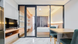 A modern hotel room with a flat-screen TV, a wooden desk, a dining table with chairs, a bed with a white mattress, and a sliding door leading to a balcony. - Gorgeous 1Br Apartment At The Smith Alam Sutera (Tangerang)