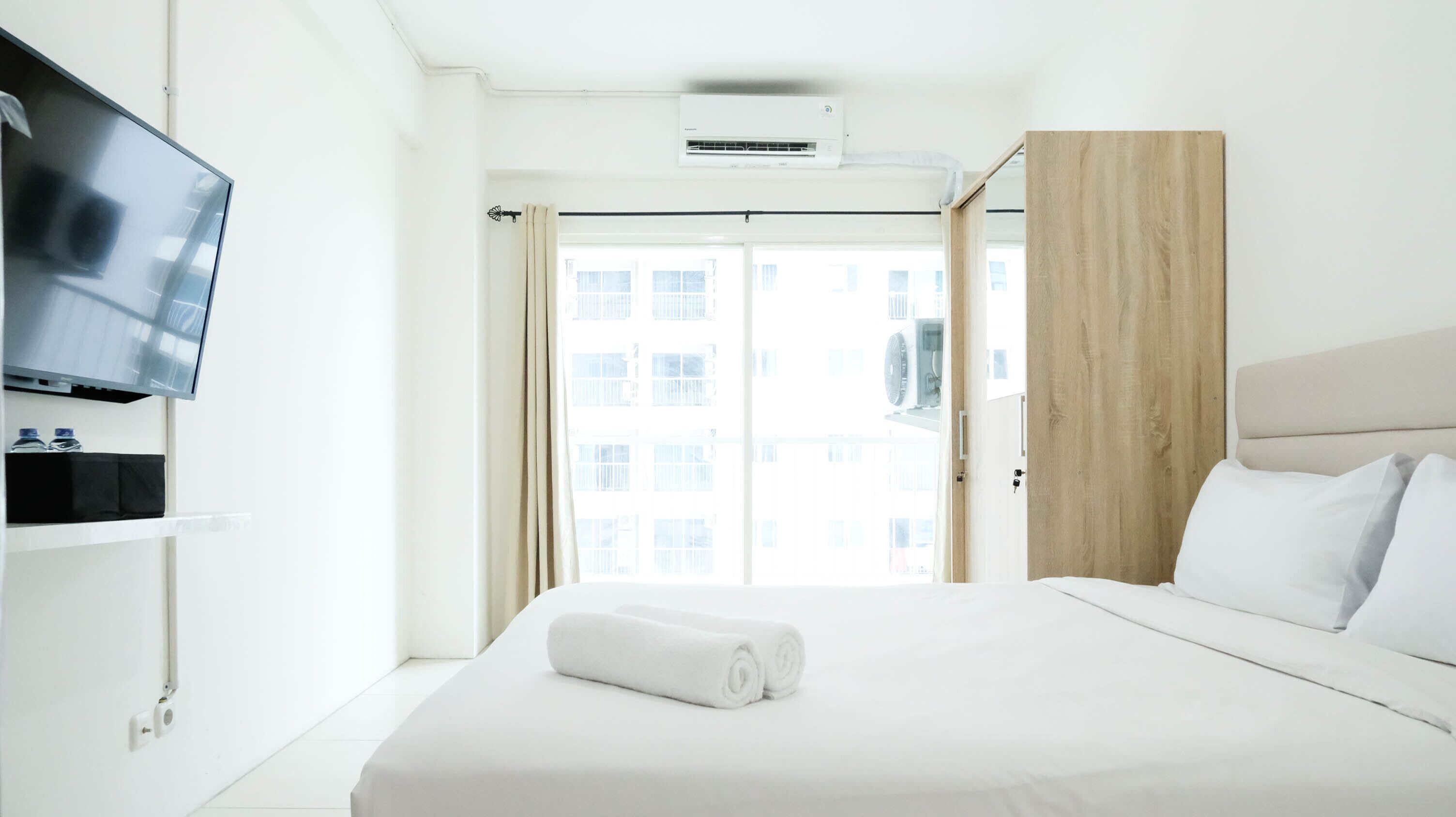 A hotel room with a bed, a television mounted on the wall, an air conditioning unit, and a view of the outside through a window.