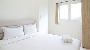 Une chambre d’hôtel propre et blanche, avec un lit, deux oreillers, une fenêtre avec des rideaux et deux serviettes enroulées posées sur le lit.