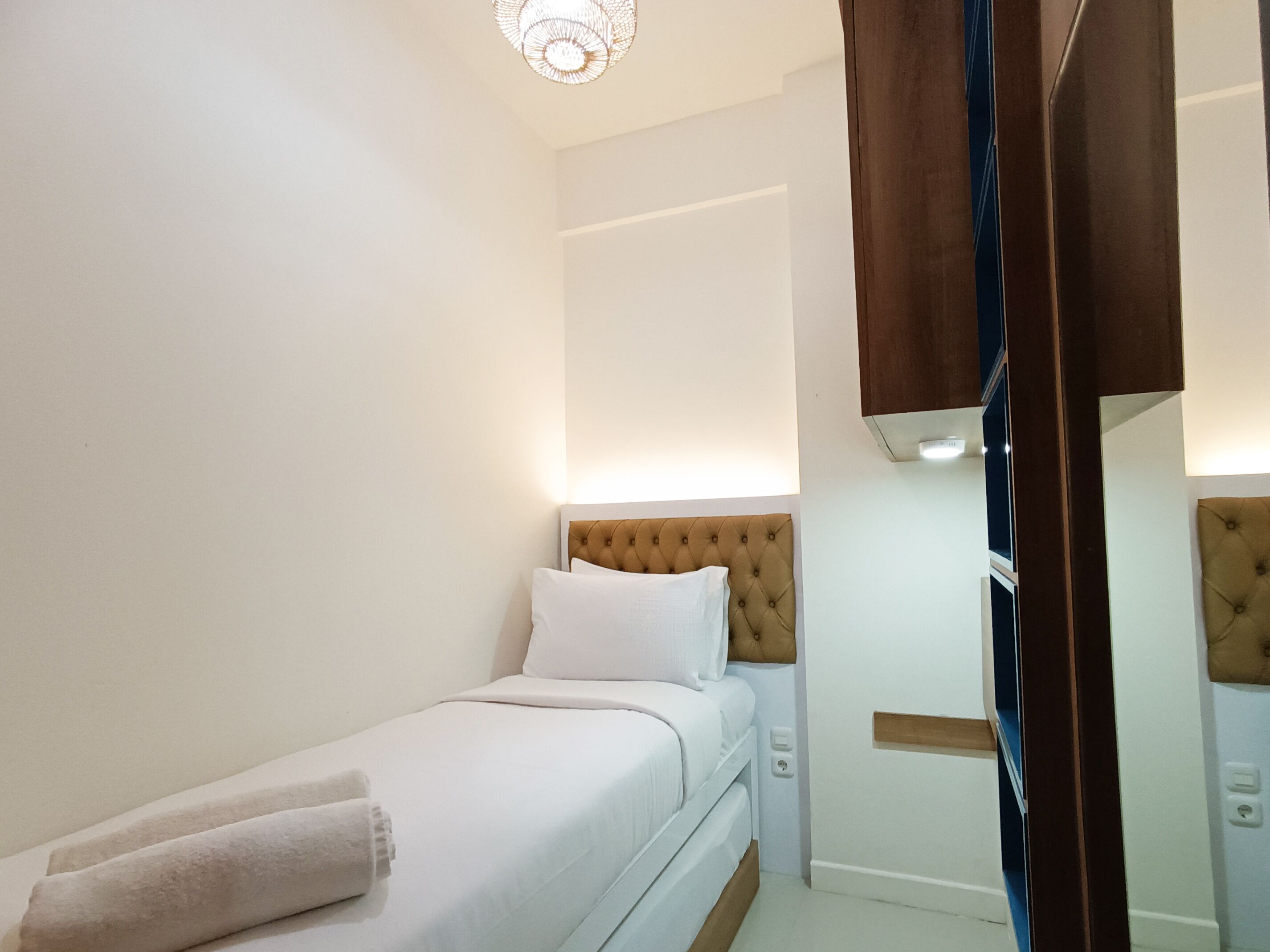 A neatly made bed with white linens and a single pillow, a bedside table, and a wall-mounted light fixture.