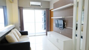 A modern living room with a sofa, a flat-screen TV mounted on a wooden wall, and a sliding glass door leading to a balcony. - Homey 2Br At Puncak Bukit Golf Surabaya Apartment (Surabaya)